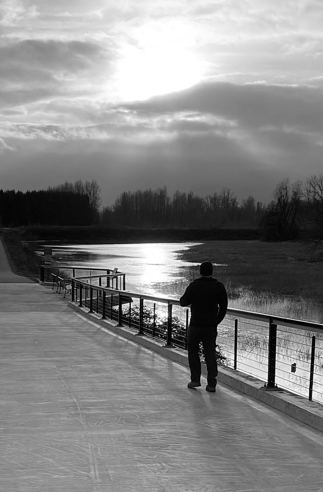 The silhouetted figure of a man walks on a paved park path overlooking a flooded meadow. The waters of the meadow reflect the diminished light of a sun partially obscured by dark clouds.