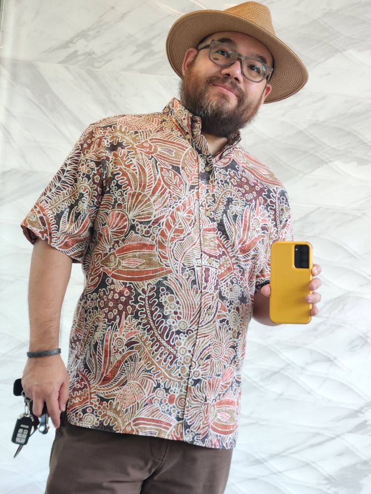Bearded man wearing a tropical shirt in orange and autumnal hues with brown trousers. He wears a paper stew hat and wood print glasses frames. He's posed in front of a textured white ceramic wall.