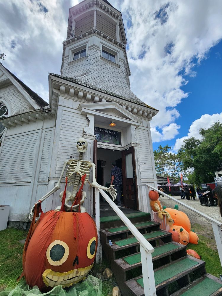 An old Victorian chapel at LA's Heritage Square Museum. It's decorated along the sides with a large skeleton sitting on a pumpkin head that looks like the character Enoch from "Over the Garden Wall." Additional pumpkins decorate the other side. 