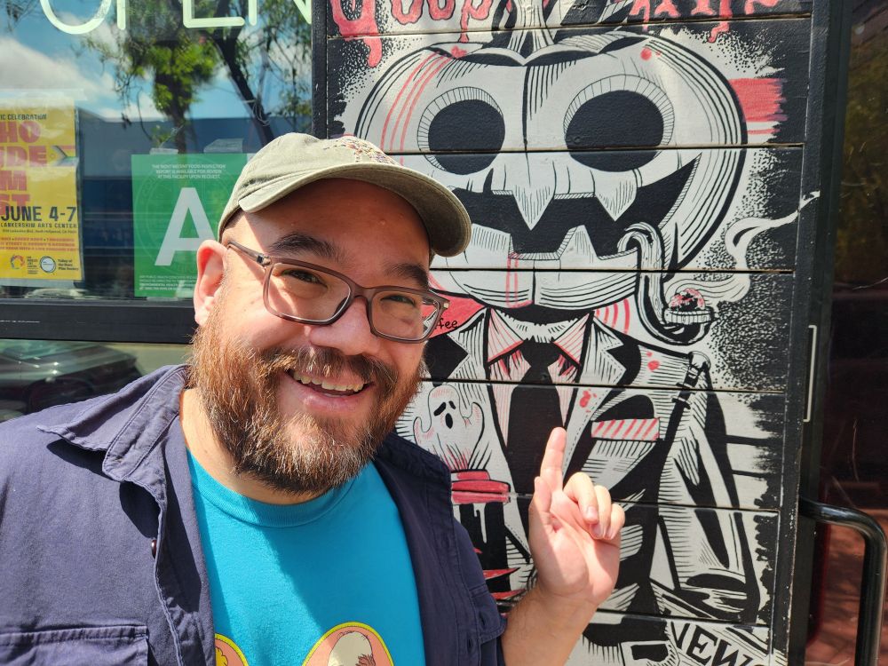 A bearded mix race man in a baseball cap poses in front of an illustrated mural of a jack-o-lantern man.