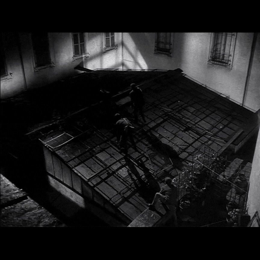 Single frame from the 1958 film Big Deal on Madonna Street. Time code: 01:28:00. Shot type: Wide/Rooftop shot of our criminals freezing like roaches a top the paneled window rooftop as the lights from indoors turns on.