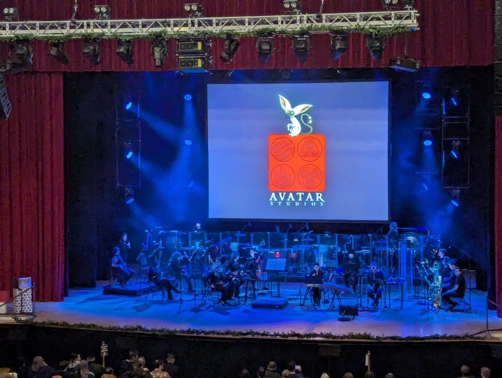 An orchestra on stage for Avatar The Last Airbender live in concert. A screen says Avatar Studios with Momo sitting on top