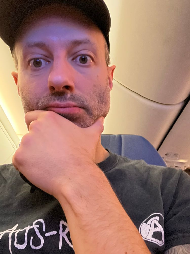 Self-portrait of a 40-something white dude who’s barely awake, sitting in an airplane seat. Wearing an Aus-Rotten t-shirt.
