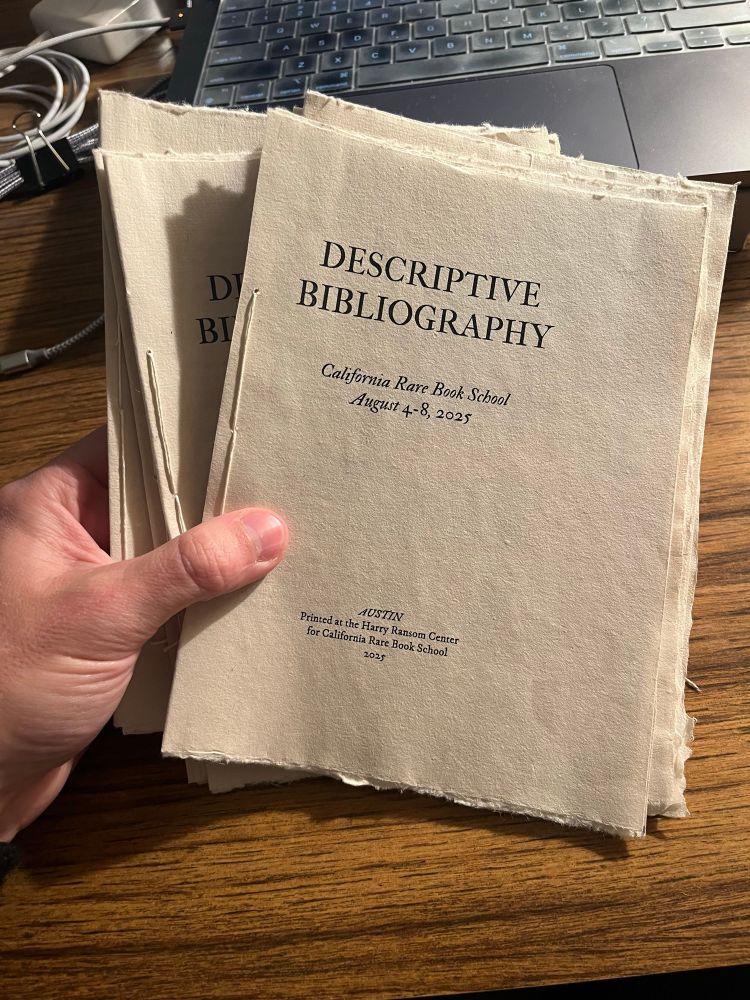 A hand gripping a stack of stab-stitched pamphlets on the surface of a table. The title page that’s visible identifies them as syllabuses for the 2025 Descriptive Bibliography course at California Rare Book School.