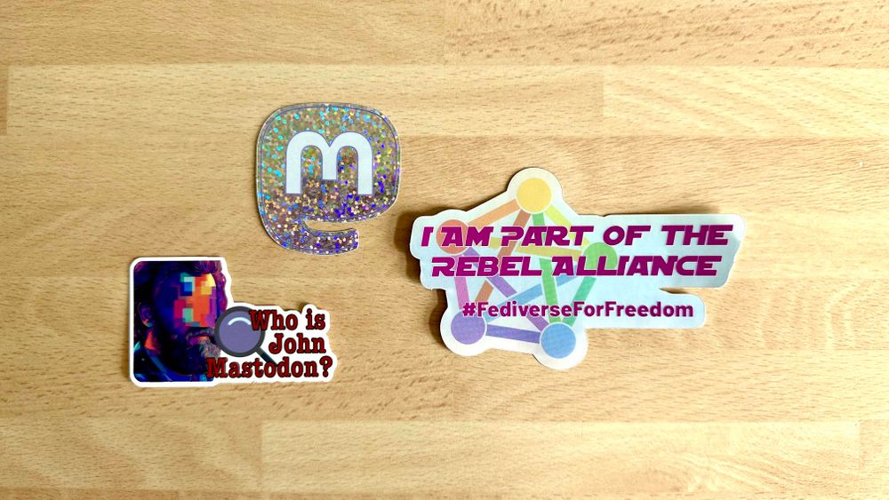 Top-down shot of three stickers: one saying “Who is John Mastodon?”, one a sparkly Mastodon mammoth logo with an “m” symbol, and the third “I am part of the rebel alliance #FediverseForFreedom”