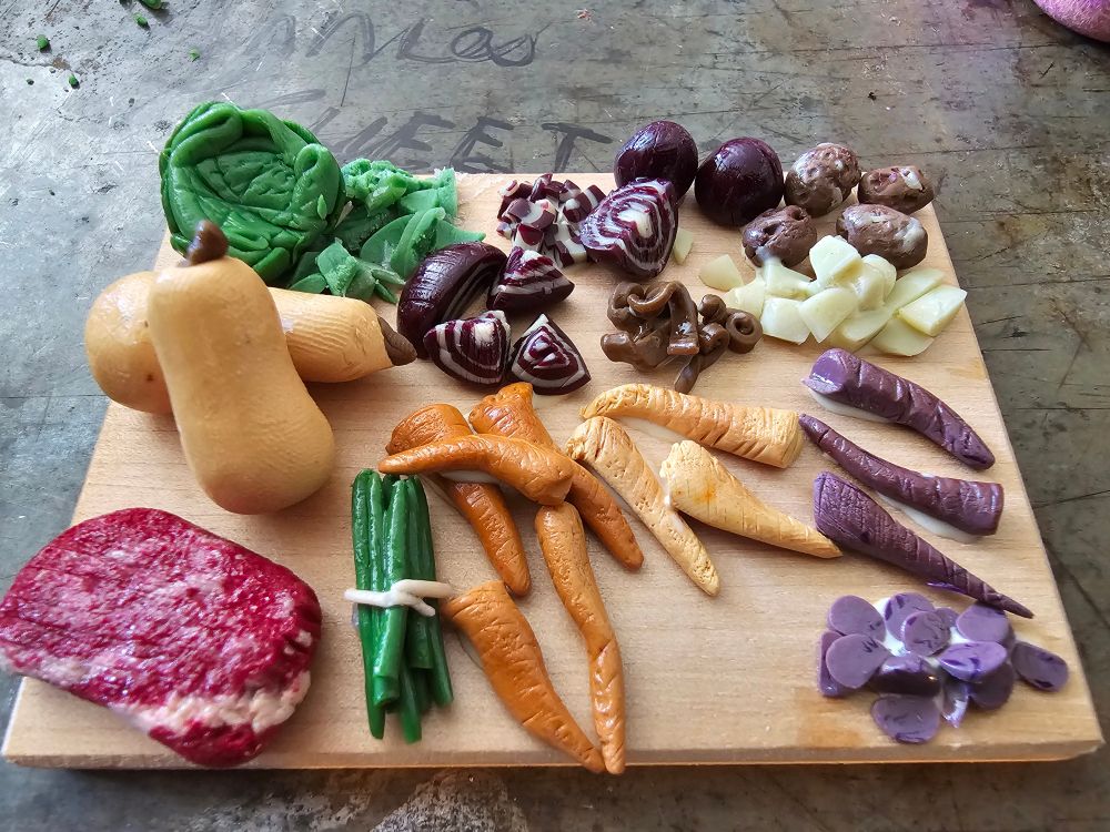 Miniature display of prepping for making a roast, with a slab of meat, asparagus, carrots of orange and purple, red onions, potatoes, apparently and butternut squash