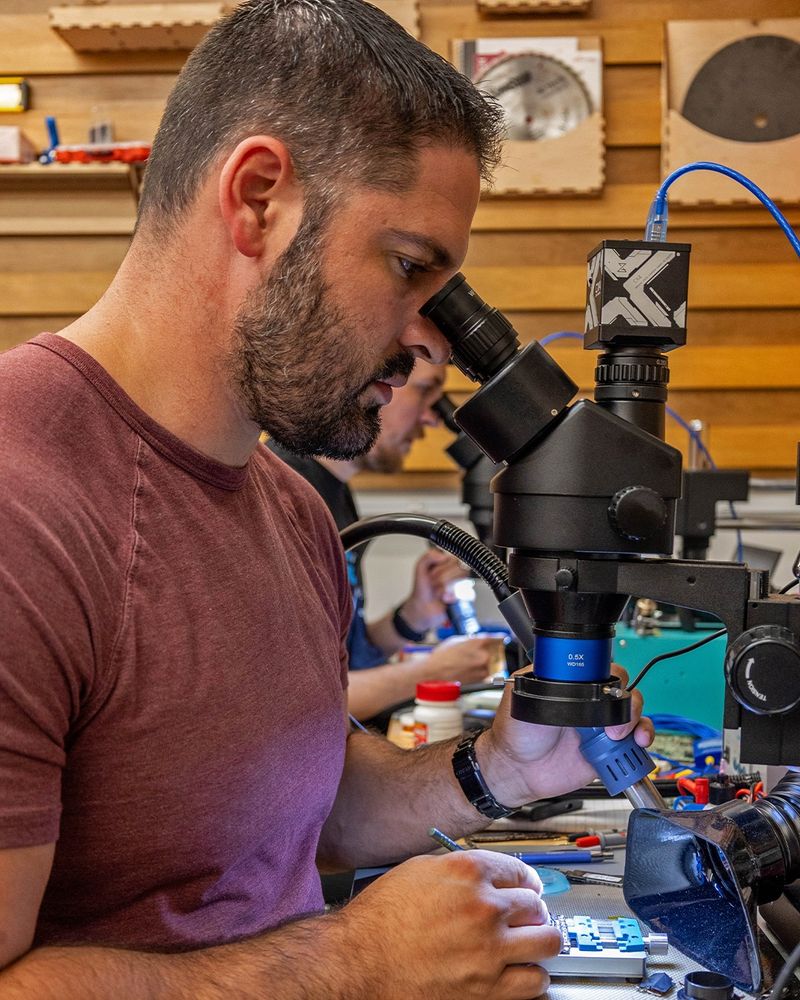A man peers into a microscope while working on a circuit board at a repair bench, surrounded by tools and electronics.