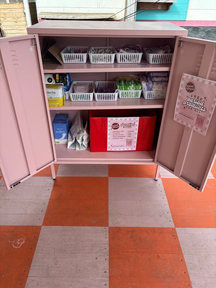A pink metal pantry with the doors open. The pantry contains period products like tampons and pads. There is a sign that reads “Free Period Products For ALL”