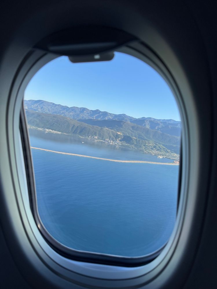Looking through an airplane window shortly after takeoff toward mountains, the edge of a city, and a causeway. Sky above the mountains is mostly blue. Much of the photo is water below the causeway.