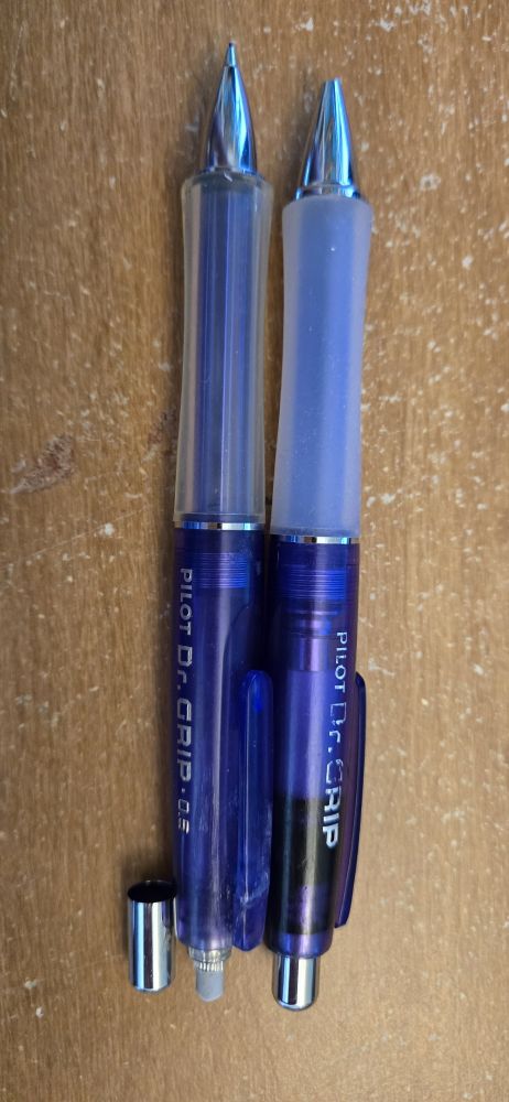 Set of purple Dr. Grips. One pen and one pencil. Pencil has cover off the eraser. Both show signs of wear and tear.