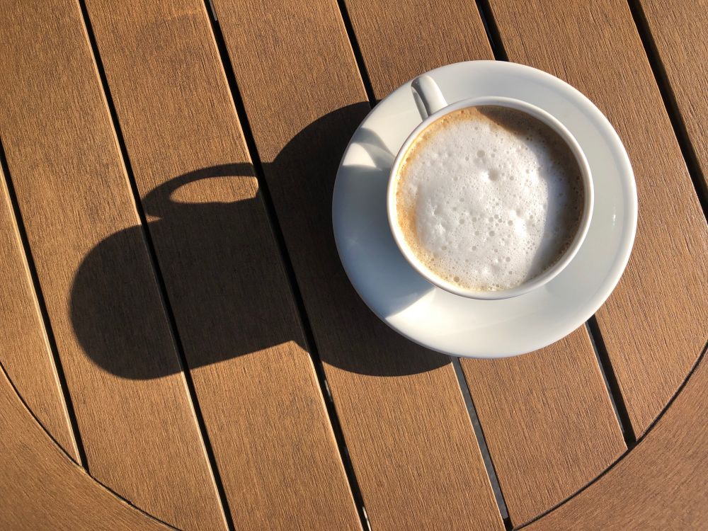 Eine Tasse Cappuccino wirft einen Schatten auf eine Tischplatte. Er zeigt quasi als Sonnenuhr in die Zeigerposition zwischen acht und neun Uhr.