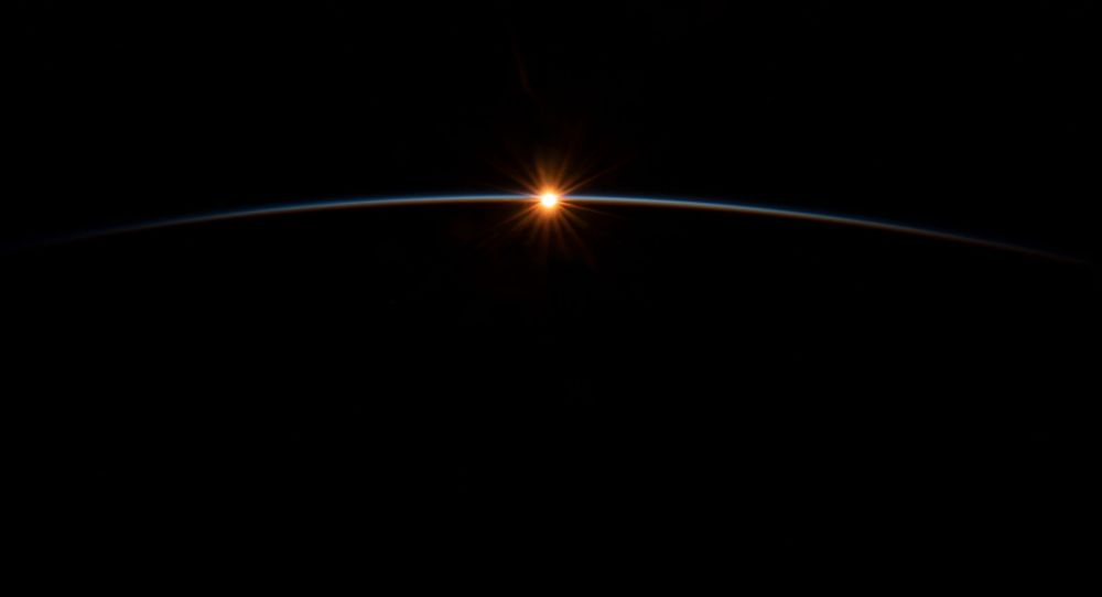 Im Schwarz des Alls in der Bildmitte die Sonne als kleiner leuchtender Punkt. Im Gegenlicht schimmert die Erdatmosphäre als dünner Streifen über der Nachtseite unseres Planeten.