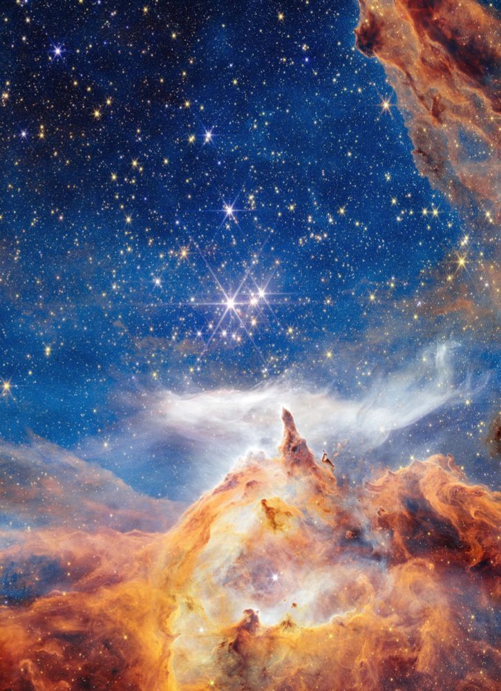 Unten erstreckt sich über ein Viertel des hochformatigen Bildes eine kosmische Wolke in warmen Farben und turbulenten Strukturen. Ein wenig davon ragt auch oben rechts an der Bildecke in die Aufnahme. Gleich aber hinzugefügt: Solche Wolken aus kleinsten Teilchen können sich zwar verdichten und so neue Sterne produzieren. Sie sind aber vor Ort zunächst mal längst nicht so massiv, wie das hier in diesem Falschfarbenbild wirkt, wo es fast wie ein "Gebirge" aus Dampf und Rauch wirkt. Darüber auf blauem Hintergrund unzählige Sterne als weiße Punkte und auch einige wenige Sterne, die von Strahlen umgeben sind - allerdings handelt es sich dabei um Artefakte, die durch die Bauweise des Teleskops verursacht werden. Vielleicht noch ein Satz zu den Farben: Es handelt sich wie oft bei solchen Infrarot-Aufnahmen um künstlich erzeugte Falschfarben, die Fachleuten unterschiedliche chemische Elemente und unterschiedliche Materiedichten anzeigen. 