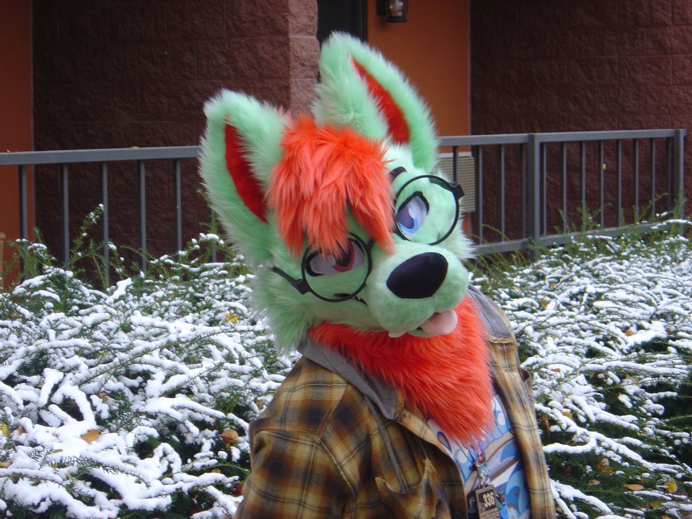 A medium close-up photo of Vidar, a green/red dire wolf fursuiter, outside the Chula Vista Resort's Bennett Tower in Wisconsin Dells. There is light snow on the bushes behind him.