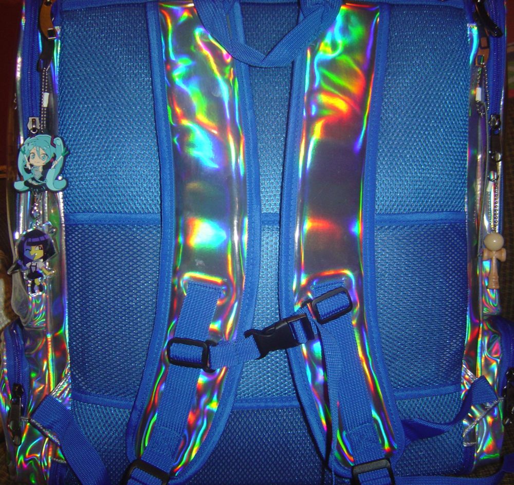 Back side of the pack, showcasing backpack straps and a suitcase slot. Attached to the zippers are a glittery acrylic ENA keychain, a Hatsune Miku ball chain charm, a Fur Squared 2025 volunteer ball chain jet tag, and a kendama ball chain charm. Each ball chain has tape over the opening.