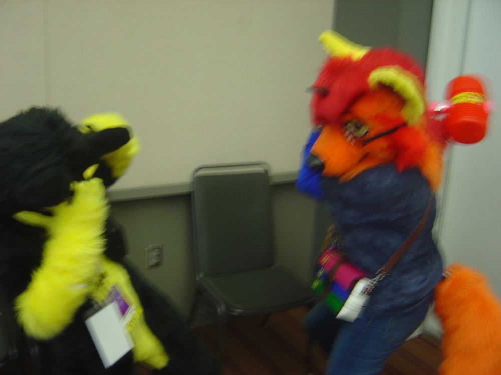 Lexicon (orange, red and yellow Tibetan fox fursuiter) wielding a red and pink squeaky hammer with Anthrocat (black and gold cat fursuiter) acting startled.