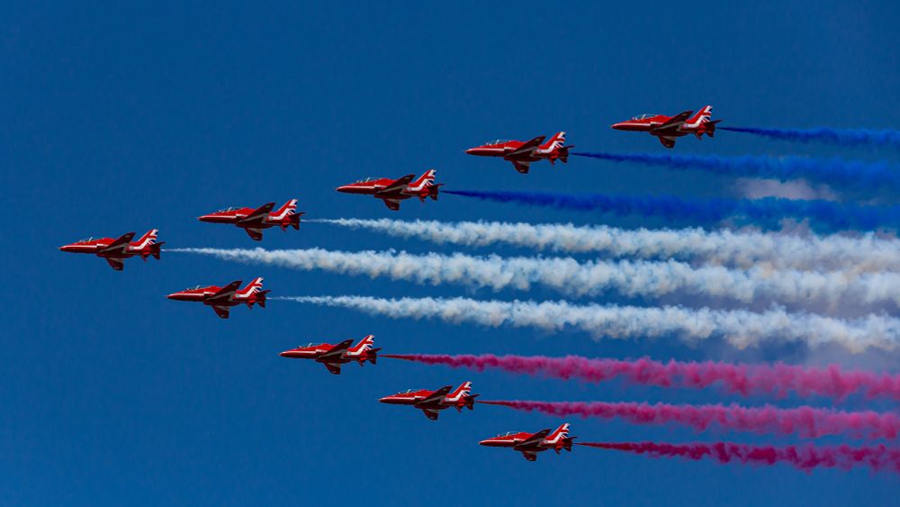 The Red Arrows flying in a V-shape formation with coloured dye smoke in Red, White & Blue across the sky