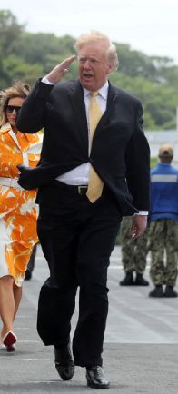 trump doing a lazy salute in a clearly windy area so he looks like an extra doofus. Wearing a yellow tie for some reason. 