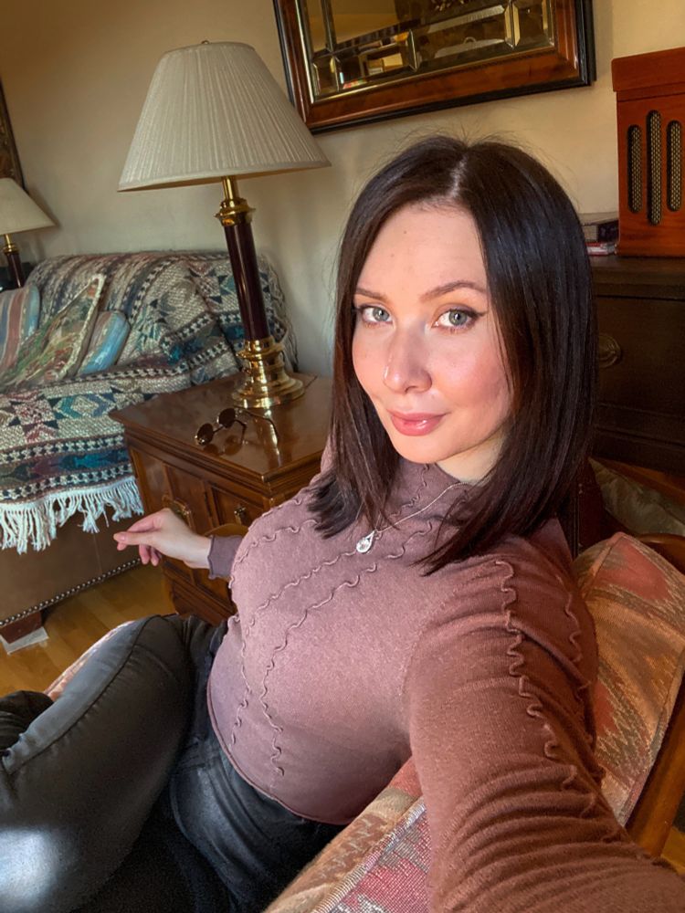 A woman with shoulder-length dark hair poses indoors, sitting on a patterned armchair. She is wearing a fitted brown sweater with decorative stitching and black pants. Her face is slightly tilted, and she gazes into the camera with a soft smile. Behind her, there is a cozy living room setup with a lamp, a wooden side table, sunglasses, and a patterned throw draped over a couch. A vintage-style radio and a mirror are visible in the background. The lighting is warm and natural, creating a relaxed and inviting atmosphere.