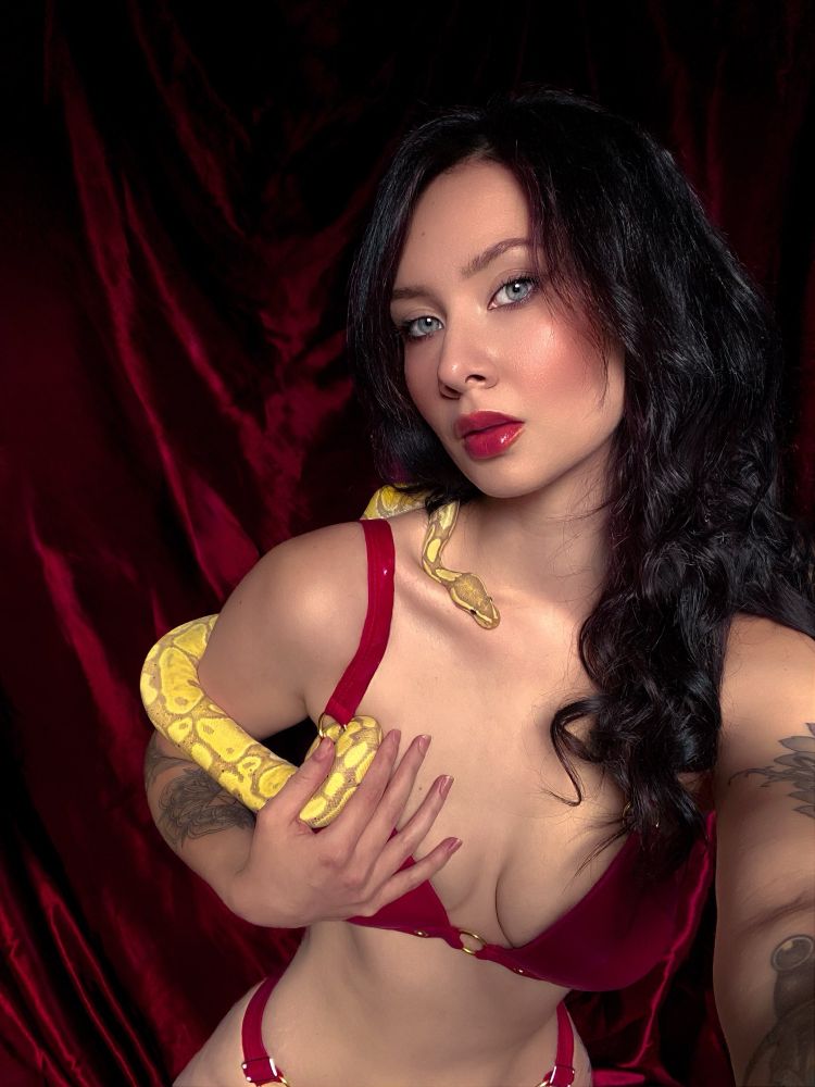 A close-up selfie of a woman with dark wavy hair and piercing blue eyes, dressed in a red leather bikini. She holds a vibrant yellow ball python draped across her chest and shoulder, set against a luxurious red velvet background. Her makeup features bold red lips, enhancing the dramatic and alluring composition.