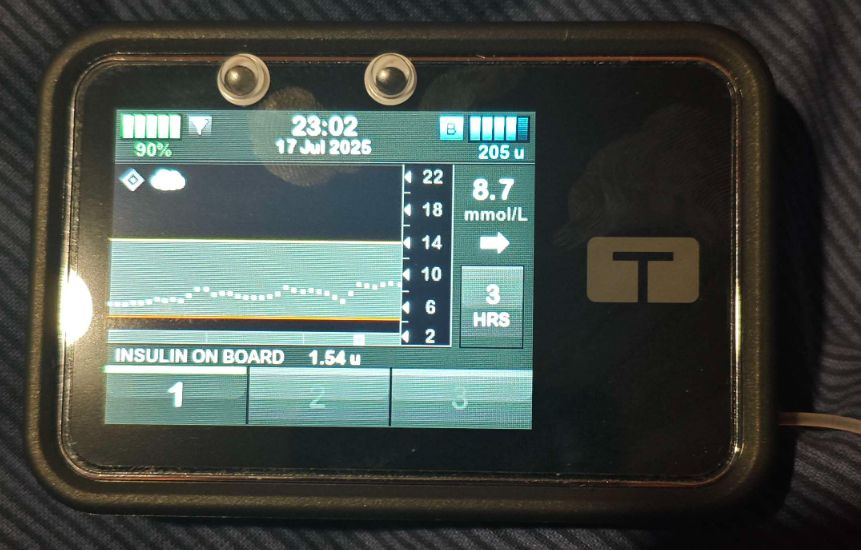 An image of a Tandem T-Slim X2 Insulin Pump. On the top, on the black part above the screen, is two googly eyes. A light is shining overhead, reflecting off the left of the pumps shiny surface. There are visible fingerprint marks on the touchscreen.