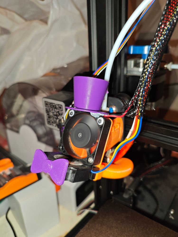 An image of a 3D printer hotend. It is orange with two black fans (the top one is facing outward toward the camera, the bottom one is horizontal and facing away from the camera.) with googly eyes on the top fan. It has a plastic purple top hat and bowtie. Various wires are coming out of the hotend, coloured blue, red, yellow and white. These wires are separated into two strips, one on the right and on the left. There is also a pair of black and red wires on the right. The background is cluttered with various things and out of focus.