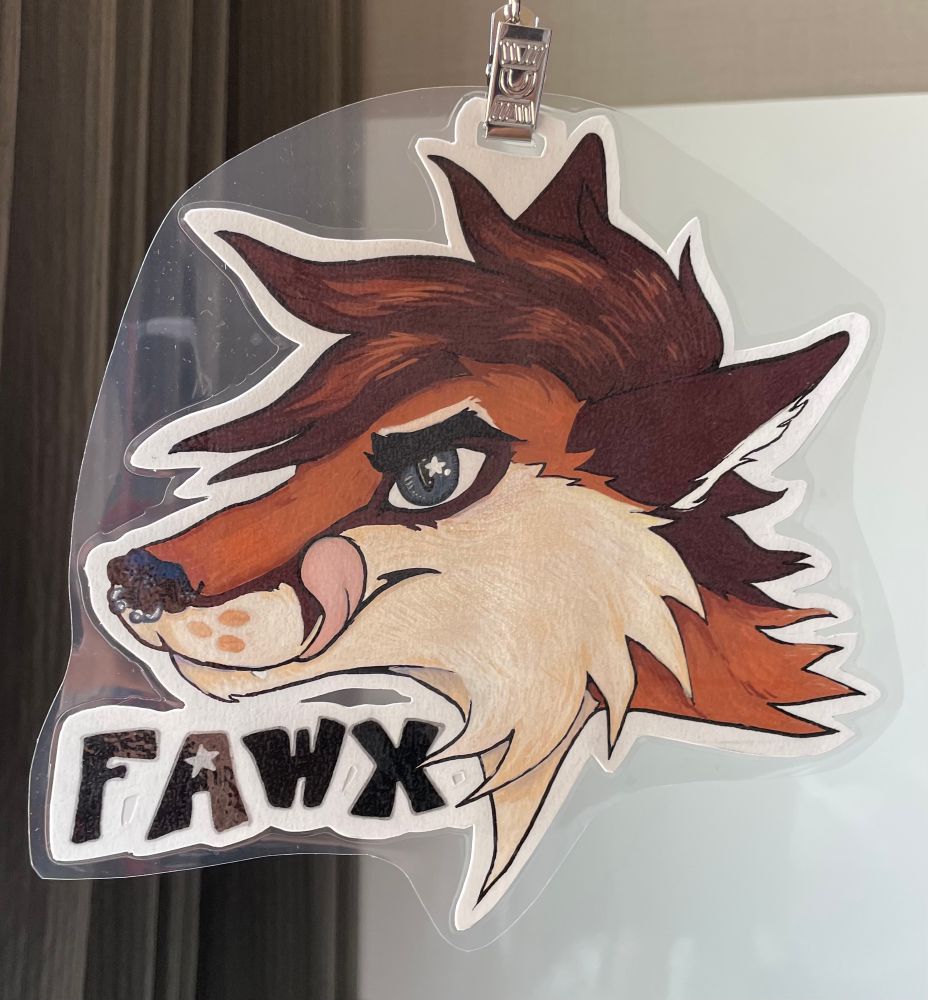 A traditionally drawn furry convention badge. It’s a headshot of a character named Fawx (text in bold black). He is brown with cream face and cheek markings. A dark brown line runs from either side of his dark nose up the muzzle and around the grey eyes. His dark brown ears are pointed back and he’s licking his lips with a pink tongue. His short brown hair is highlighted. The muzzle, ears, neck and fur almost make a star shape. The badge is hanging from a lanyard in front of a beige wall with a grey curtain and white air conditioner.