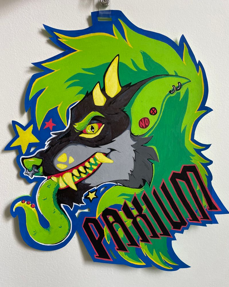 A large con badge of the alien dinodog character, Paxium. Side view looking left. He’s grinning at the viewer, showing all of his teeth and a long, bright green tongue twisting out of his mouth. Hi face is grey, dark over the chin, nose and forehead. The nose and eyebrows are the same bright green as the tongue. Three large muzzle spots, two short horns and an under eye marking are a lighter lime green. He has large bright green hair flowing over his forehead and down the right side of the badge. He has a grey spiked septum, two hoops high on his ear, and two round, red stylized earrings farther down. “PAXIUM” is written in black with a red outline at the bottom right of the badge. There are three stars around his face, two light lime green and one red.