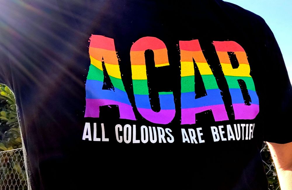 Ein schwarzes T-Shirt mit dem Schriftzug „ACAB" in großen regenbogenfarbenen Buchstaben. Darunter etwas kleiner in weißer Schrift „ALL COLOURS ARE BEAUTIFUL"