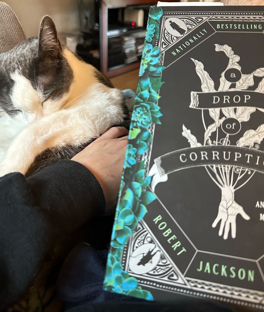 Sitting on the couch with a cat and a copy of “A Drop of Corruption” by Robert Jackson Bennett