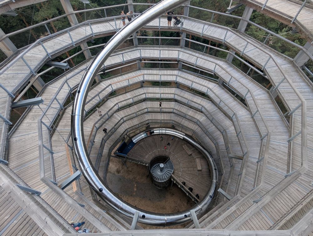 Blick von oben in das Innere eines spiralförmigen aufganges eines ca 30 Meter hohen Aussichtsturmes. Der Durchmesser beträgt ca. 10m. Von der obersten Plattform verläuft eine Röhrenrutschbahn aus Edelstahl ebenfalls spiralförmig und beschreibt in der Perspektive die Zahl Sechs. 