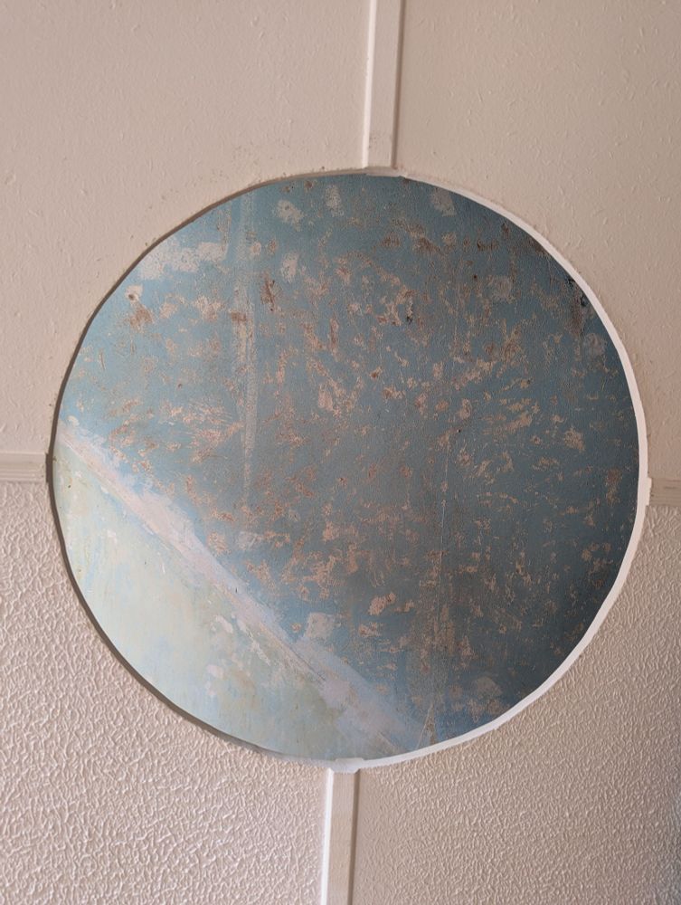 Weiße Wand mit bildfüllendem, kreisrundem Ausschnitt. Die weiße Wand im Vordergrund ist durch schmale weiße Latten in vier unterschiedlich strukturierte Bereiche unterteilt. 
Der kreisrunde Ausschnitt lässt den Blick auf eine weiter dahinterliegende, hellblaue Wand frei. Diese Wand ist unregelmäßig mit weißen und grauen Flecken und Streifen versehen, die durch Ausbesserungen oder das abkratzen von Tapeten entstanden sind. 
Insgesamt ersteht die Wirkung eines abstrakten Gemäldes oder eines Globus. 