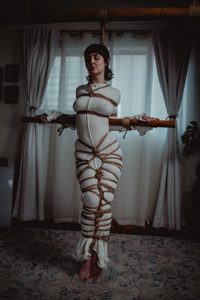 Faeriere is tied in a hauntingly lite room. They are wearing a soft form shaping white dress. Ropes are tied tightly around their legs in a shibari lacing. 

Their arms are tied to a piece if bamboo 