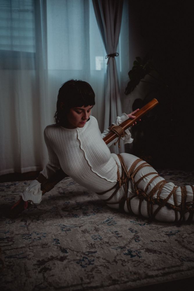 In the ground in an eerily lite space. Faeriere’s arms are tied to a piece of bamboo. Their legs are tied in a lacing pattern shibari