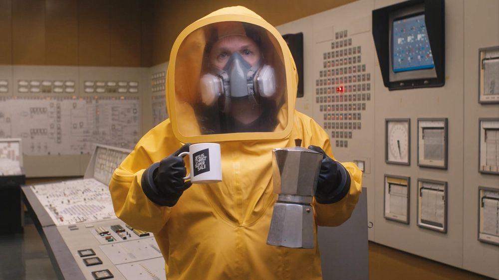 Johannes Grenzfurthner in a yellow hazmat suit inside the control room in the never-used Austrian Zwentendorf Nuclear Power Plant, holding a coffee mug and a Bialetti moka pot.