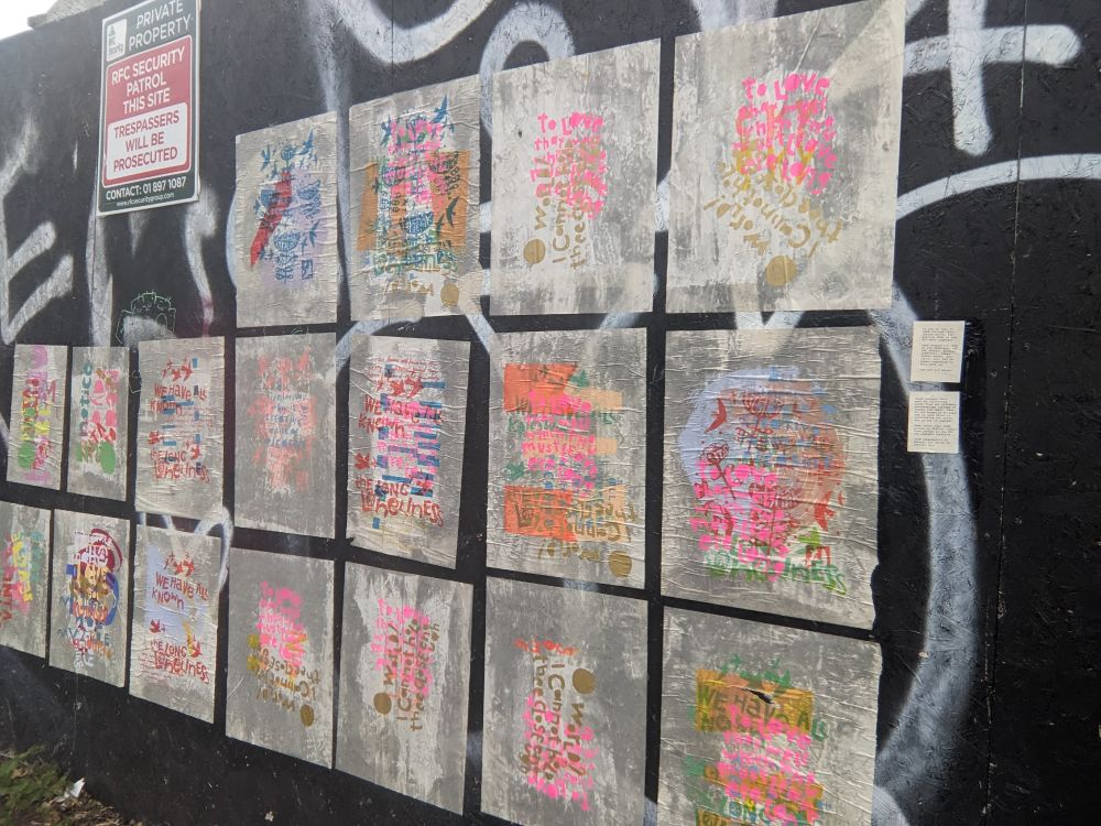 A black siding with a dozen brightly colored posters wheatpasted on it. The posters are printed with overlapping words in bright colors