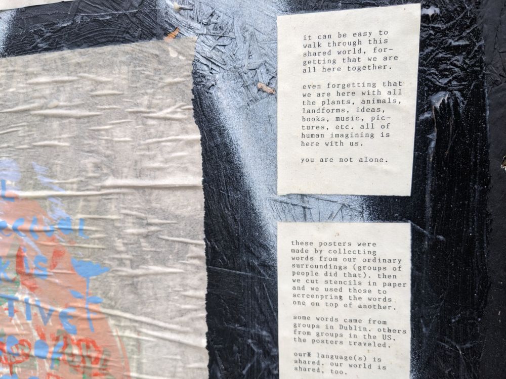 A corner of one of the posters next to small cards with typewritten language on them. The cards say, "it can be easy to walk through this shared world for getting that we are all here together. Even forgetting that we are here with all the plants, animals, landforms, ideas, books, music, pictures, etc. All of human imagining is here with us. You are not alone." and "these posters were made by collecting words from our ordinary surroundings (groups of people did that). Then we got to stencils in paper and we used those two screen print the words one on top of another. Some words came from groups in Dublin.  Others came from groups in the US. The posters travelled. Our language is shared. Our world is shared, too."