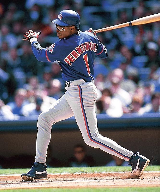 An action photo of Tony Fernandez, the infielder who played for the Toronto Blue Jays from 1983-1990, in 1993, from 1998-1999, and finally in 2001. He is wearing a blue Blue Jays jersey with his name and uniform number, 1, in red lettering with white trim. He is at home plate and has just swing his baseball bat. 