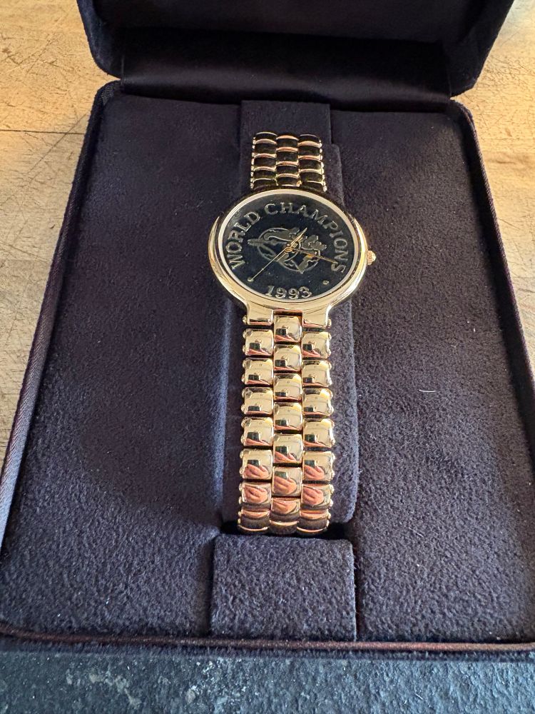 A photograph of a watch, made by Tiffany & Company, with a gold-colored band and bezel and a black face. On the face, in gold lettering, are the words “World Champions” and “1993,” surrounding a doubled image of the Toronto Blue Jays logo, signifying the back-to-back World Series victories. 