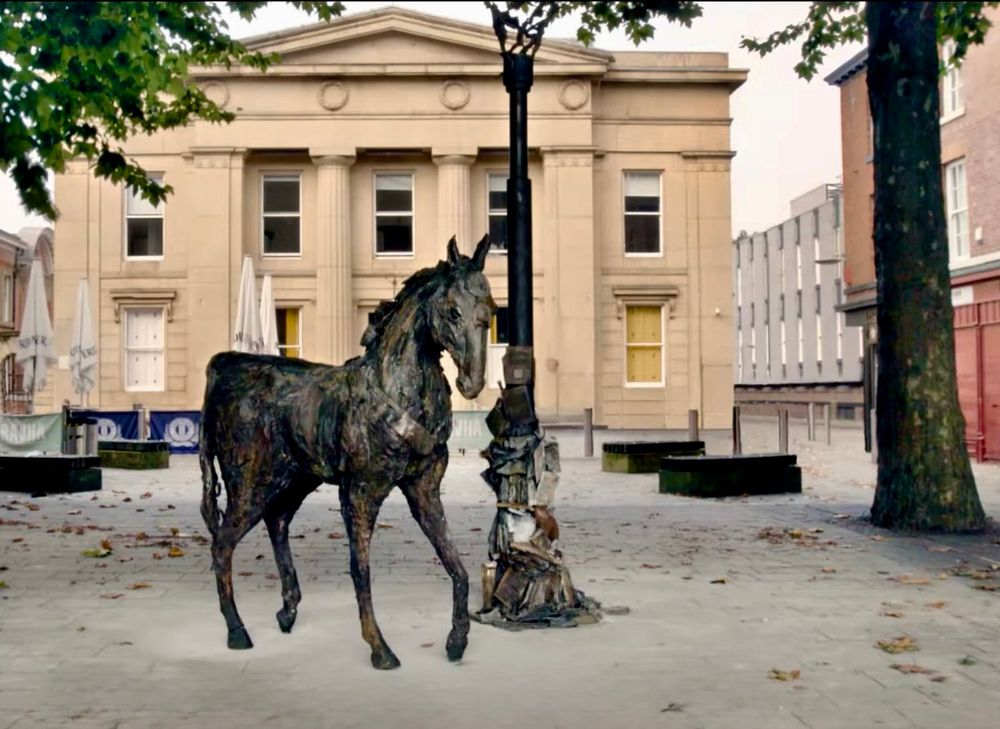 ALT text: The image depicts a public square with a horse sculpture positioned prominently in the foreground. The sculpture is crafted from a material that gives it a textured, rugged appearance. It stands adjacent to a lamp post, which is also part of the sculpture, with the horse seemingly interacting with it. The square is paved with stone tiles, and scattered leaves are visible on the ground. 

In the background, there is a neoclassical building with a symmetrical facade featuring four columns and a triangular pediment. The building has several windows, some of which appear to be boarded up. To the left of the building, there are several large umbrellas, likely belonging to an outdoor seating area. On the right side of the image, there is a tree with visible branches and leaves, and a red brick building is partially visible. The scene is set in an urban environment, with other modern buildings visible in the distance.