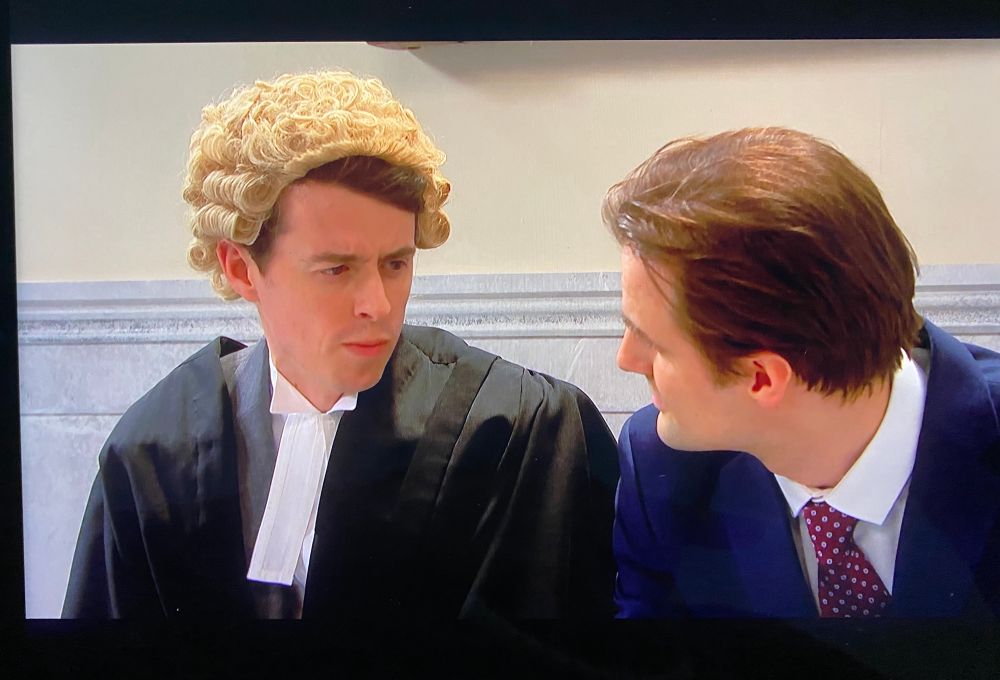 The image shows two individuals seated next to each other. The person on the left is wearing a traditional judge's wig, characterized by its curly design, and a black robe with a white neckband, typical of judicial attire. The person on the right is dressed in a formal suit, consisting of a dark jacket, a white shirt, and a tie with a pattern. Both individuals are positioned closely, suggesting they are engaged in conversation or interaction. The background features a wall with a horizontal decorative element, possibly a molding or paneling.