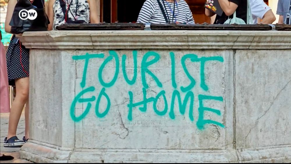 The image shows a stone structure with graffiti that reads "TOURIST GO HOME" in large, bold letters. The graffiti is prominently displayed on the front surface of the structure. Above the graffiti, there are several people standing, partially visible from the waist up. They are positioned closely together, with some wearing striped clothing and others wearing patterned tops. A black bag is visible on the left side, resting on the stone ledge. The stone structure has a detailed edge and appears to be part of a larger architectural element.