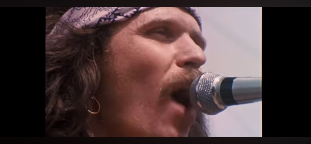 The image shows a close-up view of a microphone on the right side, positioned horizontally. The microphone has a metallic body with a mesh head, typical of standard microphones used for vocal performances or speeches. On the left side, there is a partial view of a person's hair (Country Joe), which is curly and dark brown. The hair appears to be voluminous and is partially covered by a patterned fabric, possibly a headscarf or bandana. The blurred section in the center obscures the face of the person, preventing identification or further description.