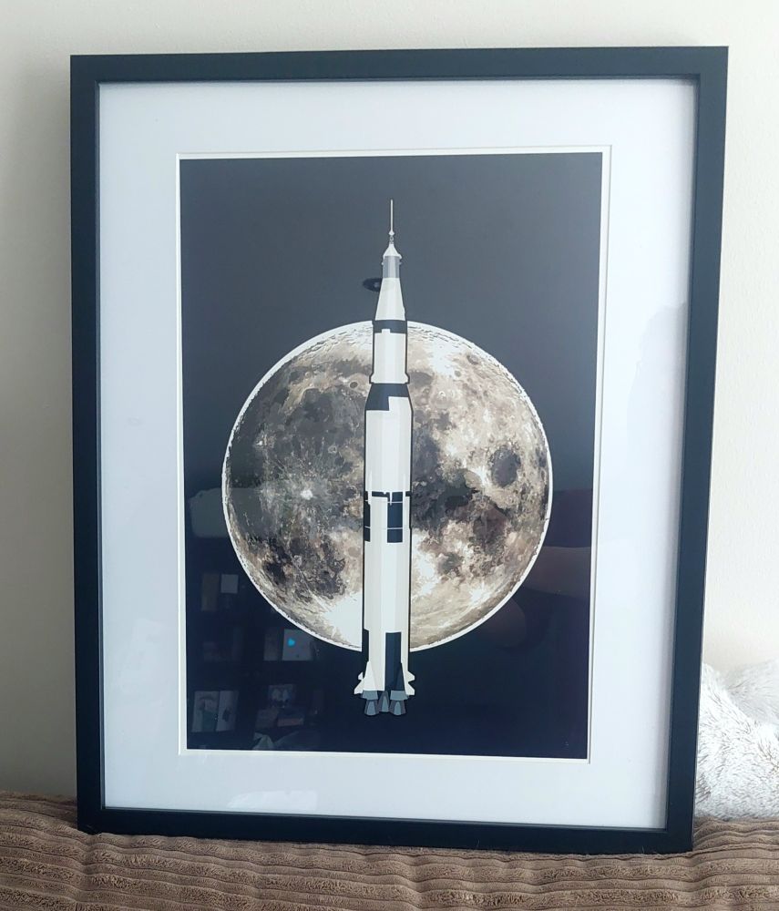 A framed print featuring the Saturn V rocket in front of the moon on a dark blue background. 