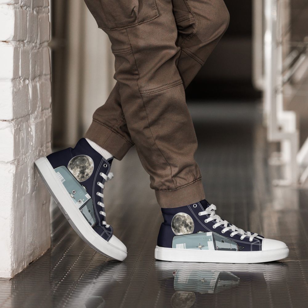 A person leaning against a wall, viewed from their feet to their upper shins. They are wearing a pair of high-top trainers made of dark blue canvass, featuring the moon and the Apollo Command & Service Module on the side panels and the Saturn V rocket on the tongues 