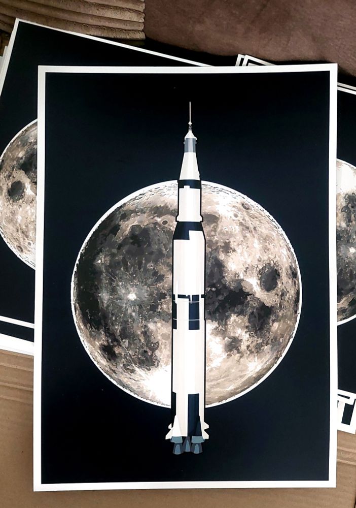 A photograph of a print of my 'moonshot' piece, a picture of the moon with the Saturn V in front of it, on a dark blue background