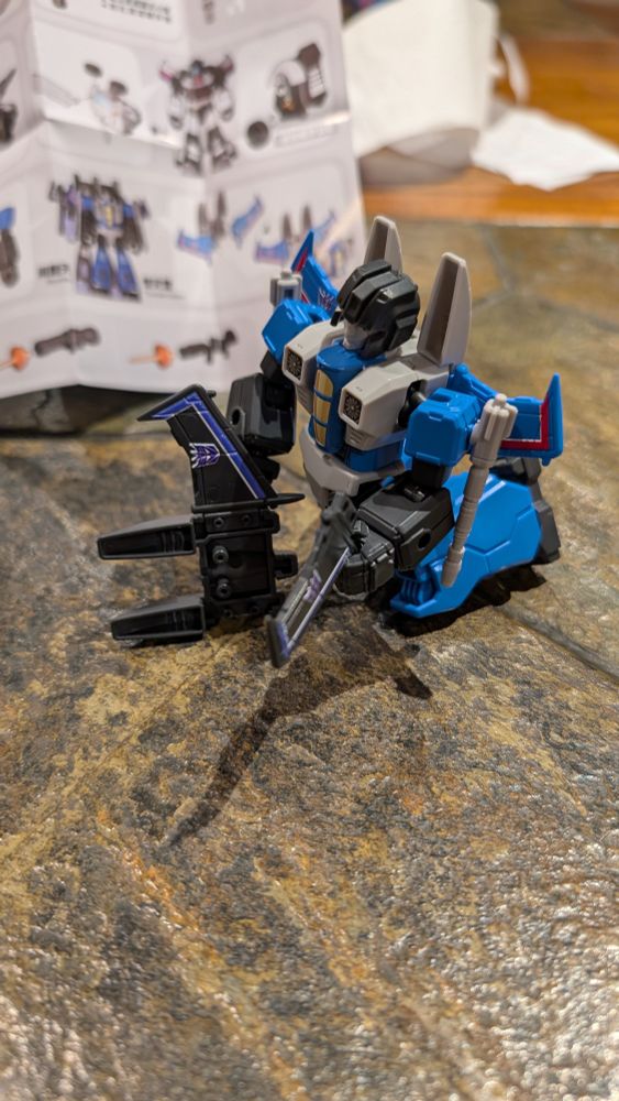 A kneeling Thundercracker assembles the wings of a Blokees Skywarp figure