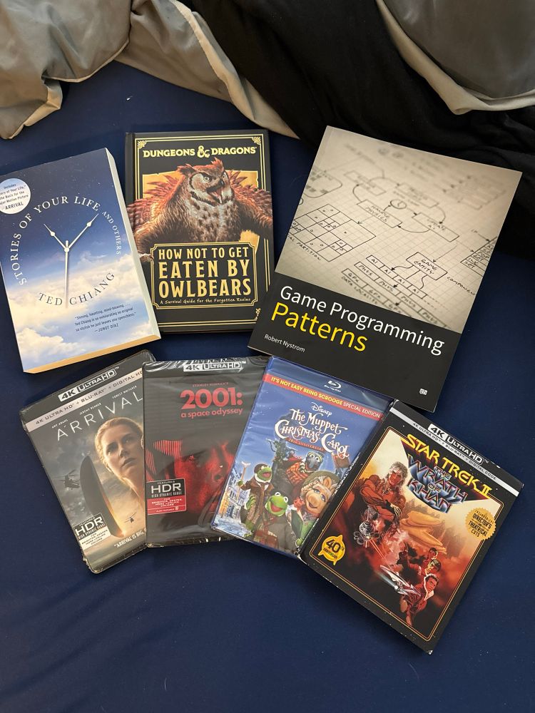 A row of books above a row for Blu-Rays. From left to right, the books are Stories of Your Life and Others, How Not to Get Eaten by Owlbears, and Game Programming Patterns. The Blu-rays are Arrival, 2001: A Space Odyssey, The Muppet Christmas Carol, and Star Trek II: The Wrath of Khan. Arrival, 2001, and Wrath of Khan are all 4K Blu-rays, and Wrath of Khan has both the theatrical and director’s cuts. The film Arrival is based of Stories of Your Life.