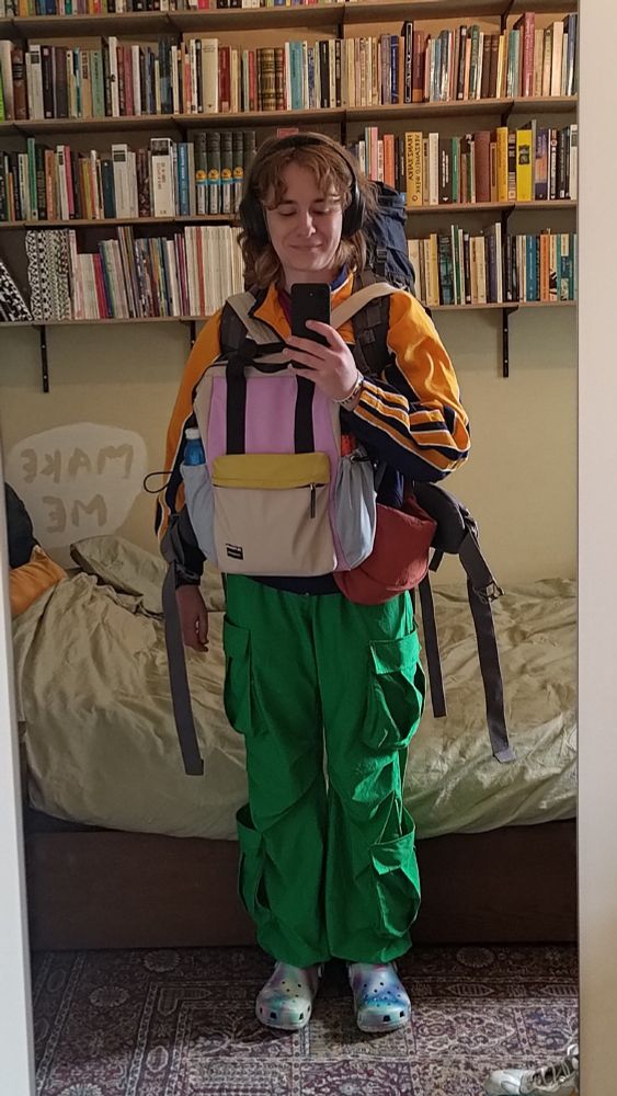 Spiegel foto van Marte die Crocs in alle kleuren draagt, een knalgroen broek, een geel jack, een oranje tasje, een rugzak voor haar buik in wit-geel-roze-blauw. Ze staat in haar slaapkamer. Achter haar haar bed en boekenplanken.