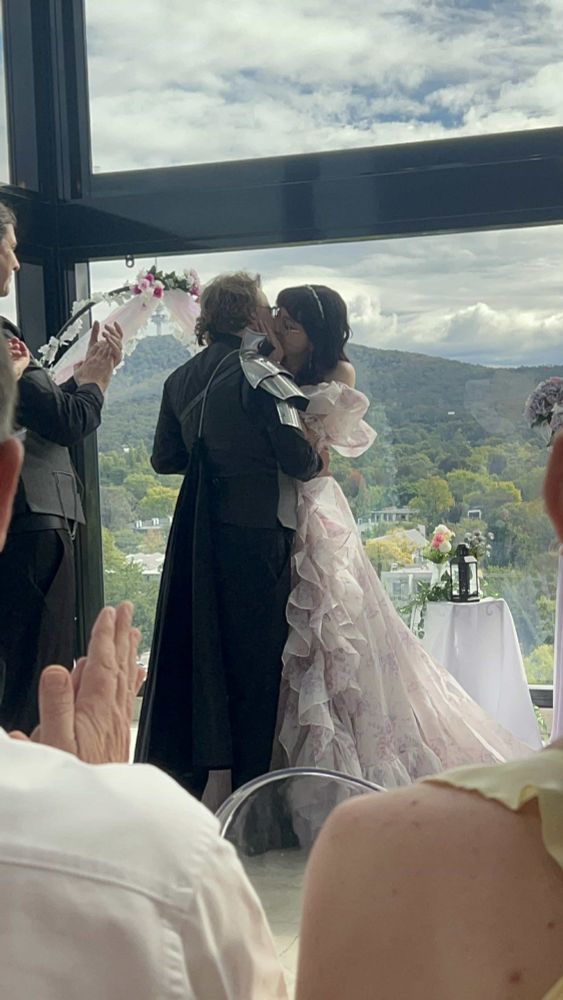 Photo from our wedding ceremony taken from the Audience, of myself (Bride) and the Groom sharing a kiss. 

On the right of the photo is myself, with a white based dress, lightly coloured with a pink and purple flower print. The dress has many ruffles, short off the shoulder puff sleeves, and a very princess like vibe.

On the left of the photo is the groom with a simple black suit, with a silver armoured plate on his right shoulder, attached to a black Cape.

Surrounding us is a beautiful view of the sky through the open windows of our venue, and many decorations like flowers, Lanterns and iridescent fabrics.

Photo cred to one of our beautiful friends who took so many photos!